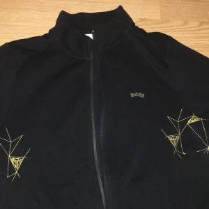 Svart vindjacka från BOSS med gul brodyr - Svart vindjacka från BOSS med coola geometriska broderier i gult på ärmarna. Jackan har dragkedja framtill och liten ståkrage. Stilren logga på bröstet och mjukt material som känns skönt mot huden. Perfekt för dig som gillar minimalistisk design med en twist. S för barn 