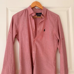 Rosa skjorta från Polo Ralph Lauren - Säljer en stilren rosa skjorta från Polo Ralph Lauren med klassisk krage och knappar framtill. Skjortan har lång ärm och broderad logga på bröstet samt broderad text på ryggen. Perfekt för dig som gillar en clean och tidlös look.