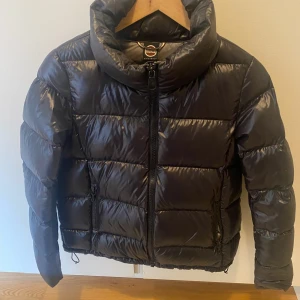 Svart pufferjacka från Colmar - Snygg svart pufferjacka från Colmar med hög krage och dragkedja framtill. Jackan har en glansig finish, två fickor med dragkedja och quiltad design. Perfekt för kalla dagar när du vill hålla stilen och värmen.