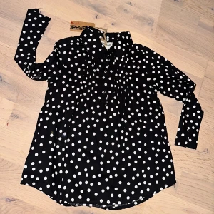 Svart prickig skjorta Barracuda Barracuda svartvit blus, storlek 86 - Svart skjorta från Barracuda med vita prickar och klassisk krage. Skjortan har knappar framtill och är långärmad. Snygg och lekfull design som sticker ut med sitt prickiga mönster.Svart och vit prickig blus med långa ärmar och krage. Blusen har knappar framtill och veckdetaljer vid axlarna.