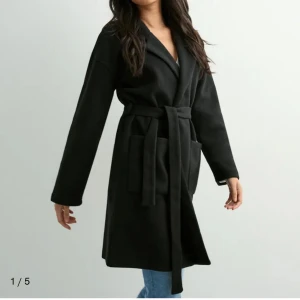 Svart kappa med bälte från nelly - Stilren svart kappa med knytbälte i midjan och stora, praktiska fickor framtill. Jackan har klassisk krage och raka, långa ärmar. Fler bilder finns