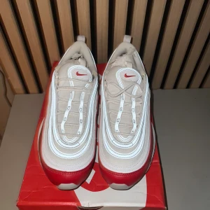 Nike Air Max 97 vit/röd sneakers - Nike Air Max 97 sneakers i en clean vit och röd färgway. Skorna har den ikoniska vågiga designen, reflekterande detaljer och synlig Air-sula. Varan är helt oanvänd och den är extremt passande för dig som gillar en blandad stil med allt ifrån Grisch till streetstyle! 