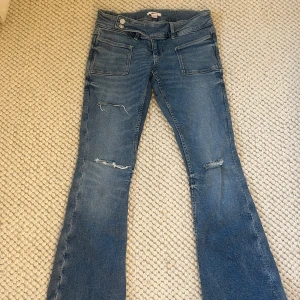 Blå bootcut jeans från Gina Tricot - Snygga blå jeans från Gina Tricot i bootcut-modell med slitna detaljer på benen och klassiska fickor fram och bak. Jeansen har två silverfärgade knappar i midjan och är tillverkade i mjukt denimtyg som ger en skön passform. Ändast annvänd fåtal gånger!!🥰