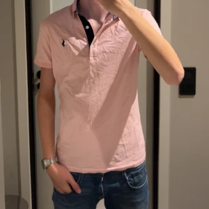 Rosa pikétröja Polo Ralph Lauren - Snygg ljusrosa pikétröja från Polo med klassisk krage och korta ärmar. Tröjan har mörkblå broderad logga på bröstet och knappar vid halsen. Tillverkad i mjuk bomull som är skön mot huden. Perfekt för en chill och stilren look. Det står storlek XXL men den passar S/M