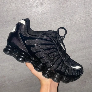 Nike Shox svarta sneakers - Nike Shox sneakers i helsvart med cool, futuristisk design och synliga fjädrande Shox-sulor. Skorna har mesh och syntet på ovansidan, snörning och diskreta detaljer i vitt och orange. Perfekt för dig som gillar sportig och edgy stil.