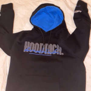 Svart och blå hoodie från Hoodrich - Svart hoodie från Hoodrich med blå insida på huvan och stor broderad logga framtill i grått och blått. Klassisk känguruficka, snörning i huvan och tryck även på ärmen och ryggen. Perfekt för dig som gillar streetwear och vill sticka ut. Price kan diskuteras ochså 