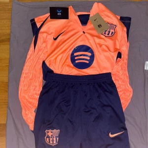 Barça långärmad fotbollströja player editon - Snygg långärmad FC Barcelona fotbollströja från Nike i orange och mörkblått. Tröjan har klubbmärke och Spotify-logga på bröstet, samt matchande byxor med FCB-emblem och Nike-logga. Tillverkad i lätt och ventilerande funktionsmaterial.