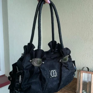 Svart axelväska med detaljer  - Cool svart axelväska från med präglat mönster, stora silvriga metalldetaljer och dubbla flätade handtag. Väskan har ett glansigt tyg och dekorativt spänne framtill med -logga. Perfekt för dig som gillar statement-accessoarer. Saknar en sådan där som man ser på sista bilden