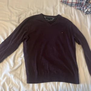 Vinröd v-ringad/lila tröja Tommy Hilfiger - Säljer en v-ringad tröja från Tommy Hilfiger i storlek L. Tröjan är tillverkad i en mjuk mix av pima bomull och kashmir, har långa ärmar och en liten broderad logga på bröstet. Perfekt för dig som gillar klassisk och stilren look.