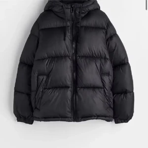 Svart pufferjacka med huva - Säljer en svart pufferjacka med huva och dragkedja framtill. Jackan har en oversized look, breda kanaler och två sidofickor. Perfekt för kalla dagar