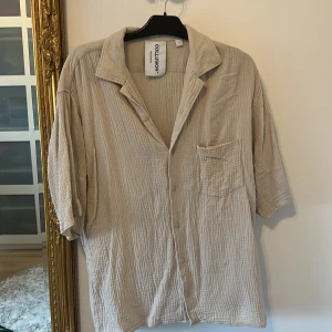 Beige skjorta från COLLUSION - Luftig beige skjorta från COLLUSION i bomull med kort ärm, avslappnad passform och bröstficka. Skjortan har en krage, knäppning framtill och ett lätt strukturerat tyg som ger en chill vibe.