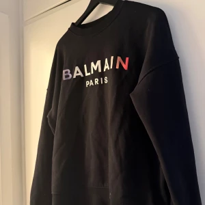 Svart sweatshirt från Balmain Paris - Svart sweatshirt från Balmain Paris med färgglad logga framtill. Tröjan har rund halsringning och långa ärmar. Tillverkad i mjuk bomull, perfekt för en clean och trendig look.