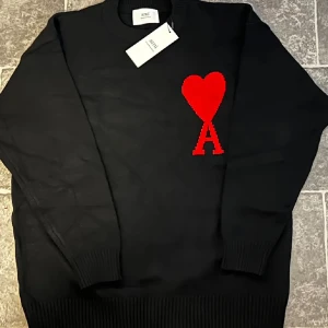Svart stickad tröja från Ami Paris - Svart stickad tröja från Ami Paris med rött hjärta och bokstaven A broderat på bröstet. Klassisk rund halsringning och ribbade muddar. Perfekt statement-plagg för dig som gillar stilrena detaljer och vill sticka ut.