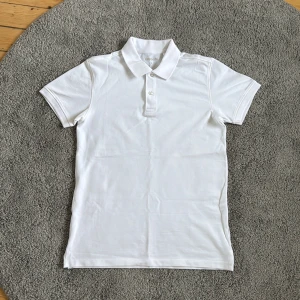 Vit pikétröja från Kappahl, storlek S - Klassisk vit pikétröja från Kappahl i storlek S. Tröjan har korta ärmar, krage och två knappar framtill. Tillverkad i mjuk bomull som känns skön mot huden. Perfekt till en clean och fräsch look.