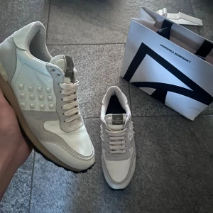 Valentino Rockrunner sneakers vita - Snygga Valentino Rockrunner sneakers i vitt med beige detaljer och coola pyramidnitar på sidorna. Skorna har snörning, rund tå och en platt sula med grovt mönster undertill. Materialmix av skinn och mocka ger en lyxig känsla och unik look.