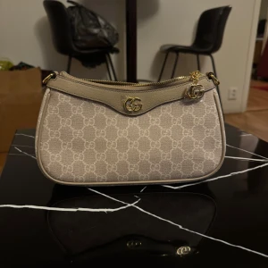 Beige axelväska från Gucci med GG-logga - Snygg beige axelväska från Gucci med klassiskt GG-monogram över hela väskan. Väskan har guldfärgade detaljer, dragkedja och medföljande bred axelrem i grönt och rött. Tillverkad i canvas och skinn, perfekt för dig som vill ha en ikonisk och trendig accessoar.