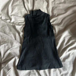 Svart ribbad polotopp ärmlös - Snygg svart ärmlös polotopp i ribbad bomull. Perfekt att styla med jeans eller kjol för en clean look. Hög hals och figurnära passform ger en modern vibe.