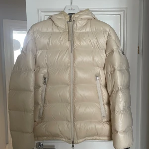 Moncler Alberic vit dunjacka, storlek 4/L  - Vit Moncler Alberic dunjacka med huva i storlek 4. Jackan har dragkedja fram och fickor med dragkedja. Detaljer med ränder i rött, vitt och marinblått finns på ärmarna och huvan. Moncler-logotypen syns på vänster ärm. Kvitto finns! Jackan är köpt på NK.