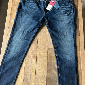 Hilfiger Denim blå jeans 31/32 - Mörkblå jeans från Hilfiger Denim med snygga slitningar och klassiska fem fickor. Jeansen har raka ben och normal passform, med tydliga kontrastsömmar och Hilfiger-logga på bakfickan. Perfekta för dig som gillar en tidlös och stilren look.