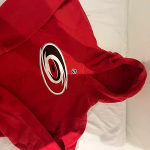 Röd NHL hoodie Carolina Hurricanes - Säljer en röd hoodie från Outerstuff med Carolina Hurricanes logga stort på bröstet och NHL-märke. Tröjan har huva och långa ärmar, perfekt för dig som gillar hockey och vill ha en sportig look. Materialet är mjukt och skönt, troligen bomull eller polyester. Köpa från NHL shop