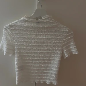 Vit ribbad croppad topp - Säljer en vit croppad topp med ribbad struktur och korta ärmar. Toppen har en lätt rynkad effekt över hela tyget och en liten volangkant vid hals, ärmslut och nederkant. Perfekt att styla med jeans eller kjol för en fräsch look.