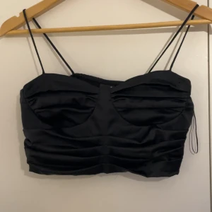 Svart satin bandeau topp - Snygg svart bandeau topp i satin med tunna axelband och veckade detaljer framtill. Perfekt för dig som vill ha en stilren och trendig look. Croppad modell som ger en modern vibe.