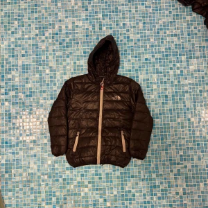 Svart dunjacka från The North Face - Svart dunjacka med huva från The North Face. Jackan har quiltad design, två sidofickor med dragkedja och ljusgrå detaljer vid dragkedjorna. Perfekt för kalla dagar och har en clean, sportig look som funkar till streetwear.