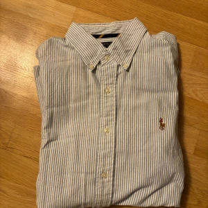 Randig skjorta från Polo Ralph Lauren XL - Klassisk skjorta från Polo Ralph Lauren i blåvitt randigt mönster. Skjortan har button-down krage, broderad logga på bröstet och knäppning framtill. Tillverkad i mjuk bomull och har långa ärmar med manschettknappar.