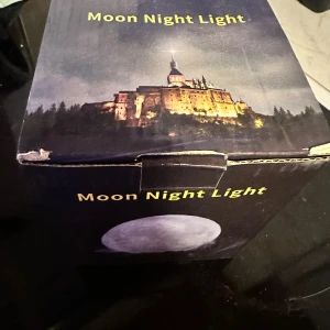 Moon Night Light Mån Lampa Nattlampa - Dekorativ nattlampa formad som månen med fjärrkontroll för färgval och ljusstyrka. Levereras med USB-kabel och träställning. Perfekt för mysig belysning i sovrum eller barnrum. Kartongen har mindre slitage men produkten ser ut att vara i gott skick.Säljer en månlampa som kan användas som nattlampa. Den kommer med en fjärrkontroll och en sladd. 🎁 