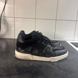 Louis Vuitton svarta sneakers i skinn - Svarta Louis Vuitton sneakers med klassiskt monogrammönster i skinn och mocka. Skorna har vit och svart sula, rund tå och snörning. Snygg kontrast mellan material och diskret LV-logga på sidan och hälen. Perfekta för dig som vill sticka ut med lyxig streetstyle.