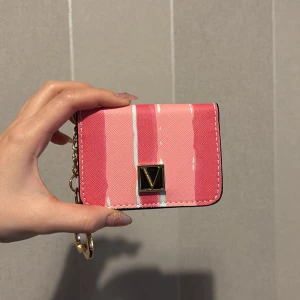 VS korthållare  - Söt liten plånbok i rosa med vita och ljusblå ränder. Plånboken har guldfärgad kedja med hjärtformat hänge och ett V-format metallmärke framtill. Insidan har plats för kort och mynt, och baksidan har ett fönster för ID eller busskort. Perfekt att ha i väskan eller hänga på nyckelringen.