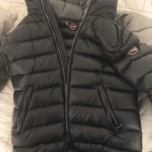 Svart pufferjacka från Colmar - Säljer en svart pufferjacka från Colmar med glansig finish och horisontella sömmar. Jackan har huva, två snedställda fickor med dragkedja och tydlig Colmar-logga på ärmen. Perfekt för kalla dagar och riktigt snygg till streetwear.