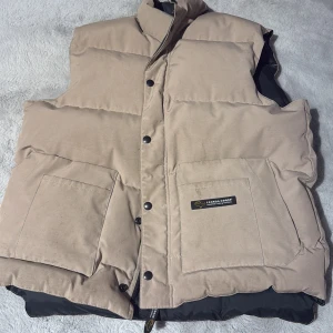 Beige dunväst från Canada Goose - Säljer en beige dunväst från Canada Goose med stora framfickor och hög krage. Västen har svarta detaljer på insidan och stängs med både dragkedja och tryckknappar. Perfekt för lager-på-lager och riktigt snygg till streetwear.