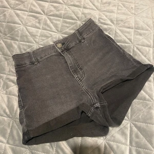Svarta jeansshorts från DIVIDED - Säljer ett par svarta jeansshorts från DIVIDED med hög midja och klassisk femficksdesign. Shortsen har knapp och dragkedja framtill, bälteshällor och uppvikta ben. Materialet är mjuk bomull med lite stretch för skön passform.