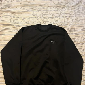 Svart sweatshirt från Prada - Svart långärmad sweatshirt från Prada med rund hals och ikonisk metalltriangel-logga på bröstet. Tröjan har en clean och stilren look med ribbade muddar vid hals och ärmslut. Materialet är mjukt och har en lätt glansig finish, vilket ger en lyxig känsla.