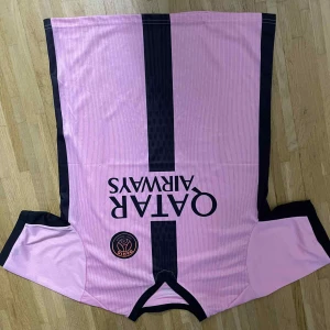 Rosa PSG fotbollströja med svarta detaljer - Snygg rosa PSG fotbollströja med svarta paneler längs sidorna och en svart vertikal rand på framsidan. Tryck med Qatar Airways och PSG-logga på bröstet. Kortärmad modell i lätt och ventilerande material, perfekt för träning eller match.