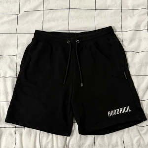 Svarta shorts från Hoodrich med snörning - Svarta shorts från Hoodrich med vit logga på vänster ben. De har elastisk midja med snörning och två sidofickor. Tillverkade i en mjuk bomulls- och polyestermix som känns skön mot huden. Perfekta för chill dagar eller träning.
