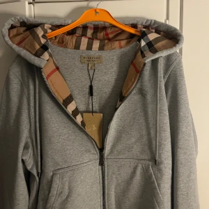Grå Burberry hoodie med rutig huva - Säljer en grå hoodie från Burberry i storlek M. Tröjan har dragkedja, klassisk rutig huva i beige, svart och röd, samt broderad Burberry-logga på bröstet. Mjuk bomullskvalitet och praktiska fickor framtill. Perfekt för dig som gillar stilrena och lyxiga detaljer.