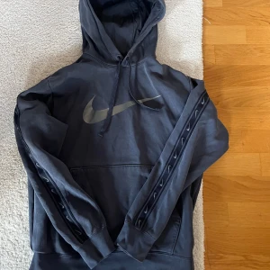 Svart Nike hoodie med logga, strl S - Svart hoodie från Nike i storlek S med stor silvrig swoosh-logga på bröstet och mindre loggor längs ärmarna. Tröjan har huva med snörning och en klassisk magficka. Perfekt för dig som gillar sportig och avslappnad stil. Passar for M. Jag köpa från JD sports 80€