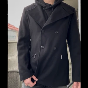 Svart Peacoat  - Tja! Säljer en svart Peacoat med dubbelknäppning. Jackan har två fickor med dragkedja och en stilren, klassisk look. Den passar dig med storlek S/M. Perfekt för dig som vill ha en snygg och tidlös jacka med coola detaljer, perfekt nu inför hösten!