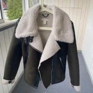 Mörkgrön teddyjacka med dragkedja - Snygg mörkgrön teddyjacka från H&M med stora kragar och mjukt, vitt foder. Jackan har coola detaljer i svart skinnimitation och dragkedjor framtill. Perfekt för dig som gillar en chill och trendig look med lite edge.