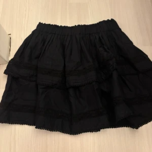 Svart kort volangkjol med spets från BikBok - Supersöt svart kortkjol från BikBok med volanger och fina spetsdetaljer. Kjolen har resår i midjan och broderad längs nederkanten för extra detalj. Perfekt för dig som gillar en feminin och lekfull stil. Pris kan diskuteras!