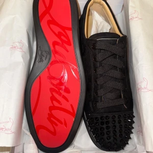 Christian Louboutin svarta skor - Svarta sneakers från Christian Louboutin med ikonisk röd sula och svarta nitar på tån. Skorna har snörning och är tillverkade i mocka med beige innersula. Perfekt för dig som vill sticka ut med en edgy och lyxig stil.