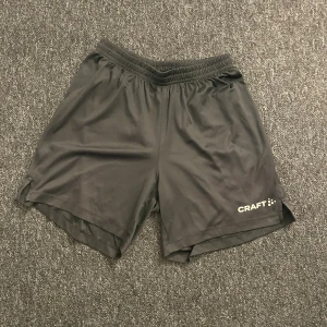 Svarta träningsshorts från Craft - Svarta träningsshorts från Craft i storlek S. De har elastisk midja och är gjorda i ett lätt och smidigt syntetmaterial som andas. Perfekta för sport och aktivitet, med diskret logga på benet och enkel design utan extra detaljer.