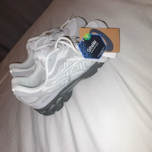 Vita och grå Asics sneakers - Fräscha Asics sneakers i vitt och ljusgrått med snygga detaljer och logga på sidan. Skorna har snörning, platt sula och meshpartier för extra ventilation. Perfekt för dig som gillar sportig stil och vill ha bekväma skor med modern look.