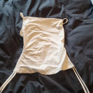 Vit linnetopp med snörning i sidan - Säljer en stilren vit linnetopp med smala axelband och snygg snörningsdetalj i sidan. Toppen är ärmlös och har en enkel, rak passform som är lätt att matcha till olika outfits. Perfekt för varma dagar och enkel att styla med jeans eller kjol.