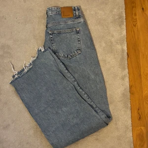  wide jeans med rå kant - Säljer ett par klassiska jeans i ljusblå tvätt med breda ben och rå, fransig kant nedtill. Modellen har fem fickor, knappgylf och en snygg vintage-vibe. Perfekt för dig som gillar avslappnad och trendig stil.