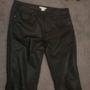 Svarta coated jeans från H&M i skinn - Snygga svarta coated jeans från H&M med fem fickor och klassisk jeansknäppning. Byxorna har en glansig finish som ger en cool look och passar perfekt till sneakers eller boots. Modellen är rak och midjehög.
