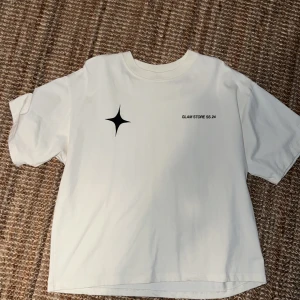 Vit t-shirt Glam Store SS24 M - Vit t-shirt från Glam Store SS24 med svart tryck fram och bak. Stor svart stjärnformad detalj och texten 'GLAM STORE FOR FAM SS 24'. T-shirten har en loose passform och är gjord i mjuk bomull. Perfekt för dig som gillar streetwear och grafiska prints. Lite Cropped men väldigt snygg och skön
