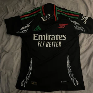 Arsenal svart bortatröja Adidas - Snygg svart Arsenal bortatröja från Adidas med gröna och röda detaljer på axlarna, vita tryck och grafiska mönster längs sidorna. Klassisk klubbkanon på bröstet och Emirates Fly Better. Helt ny